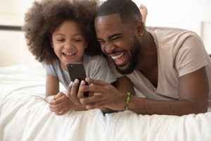 Uma criança e estudante secundarista com o seu pai. Ambos estão felizes olhando para o Joov, o melhor comparador de planos de saúde, no celular.