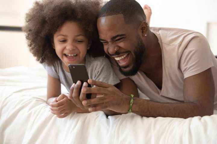 Uma criança e estudante secundarista com o seu pai. Ambos estão felizes olhando para o Joov, o melhor comparador de planos de saúde, no celular.