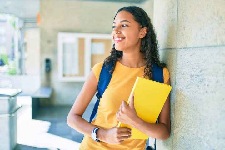 Uma estudante feliz olhando para o lado, com uma mochila nas costas e um caderno na mão.